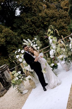 Wedding couple poses on gravel path surrounded by elegant white floral arrangements and greenery at outdoor venue.
