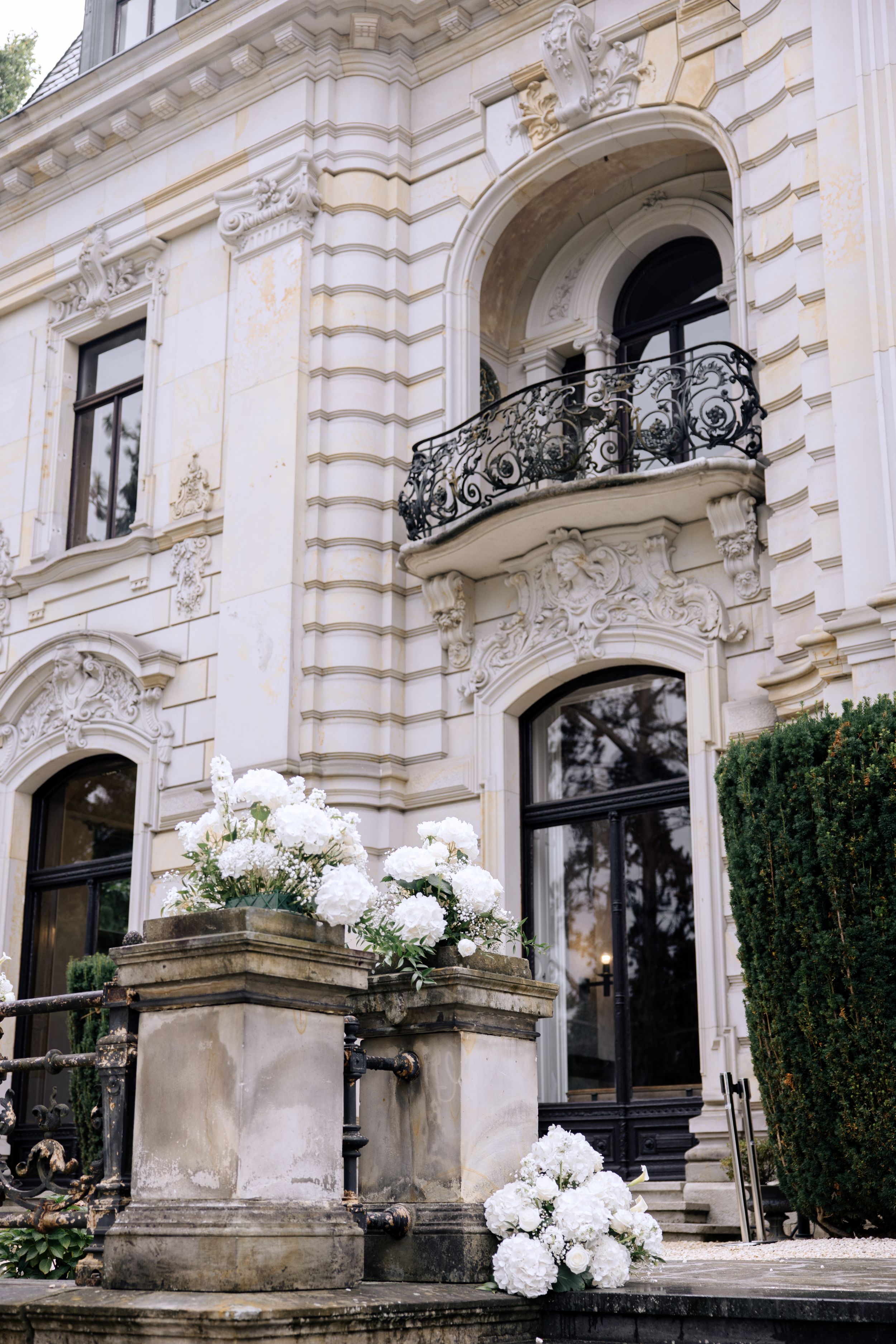 Ornate exterior entrance of a luxury building with white stone architecture and wrought iron balconies features white floral decor.