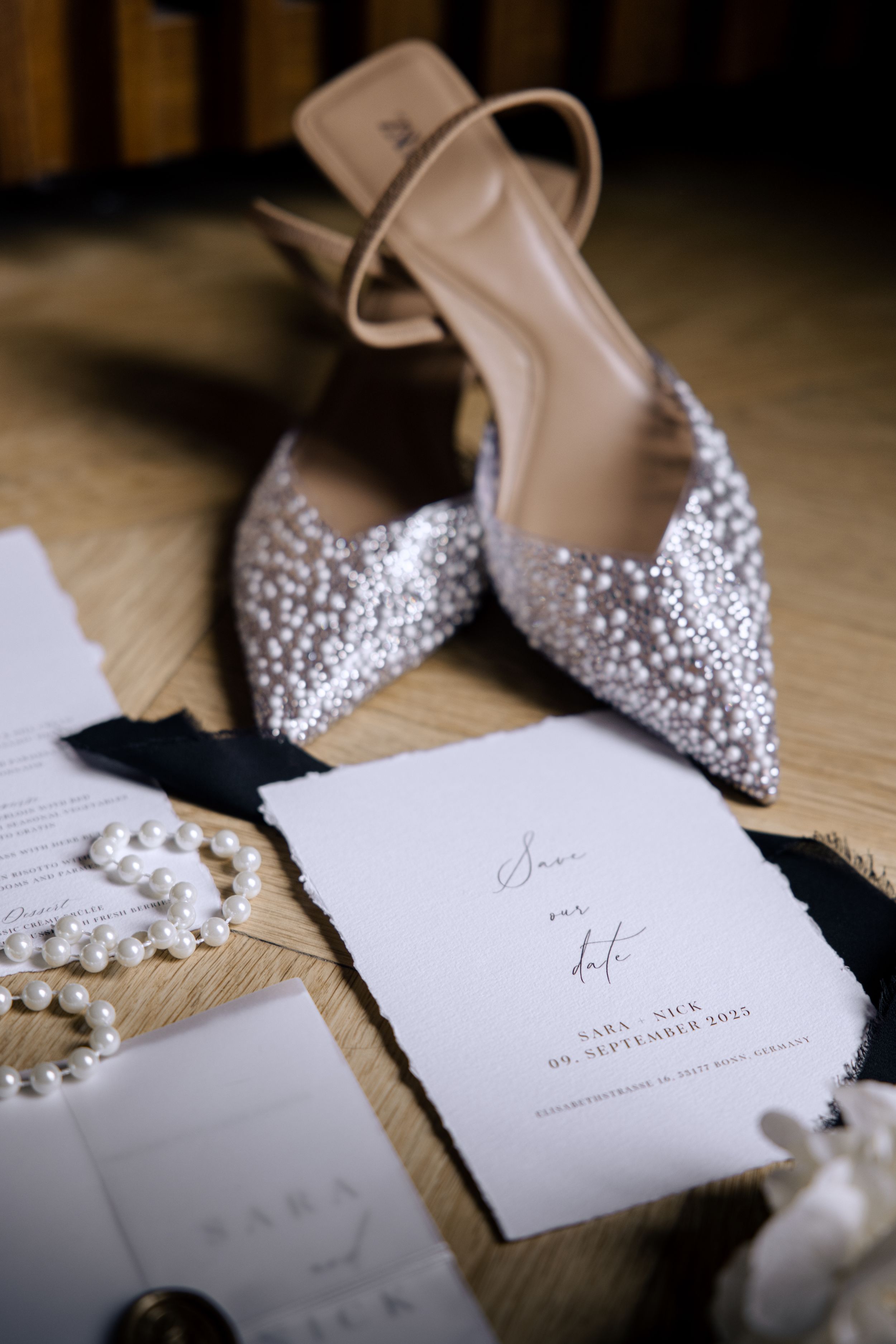 Sequence of elegant bridal shoes with crystal embellishments alongside pearl jewelry and wedding stationery on wooden surface.