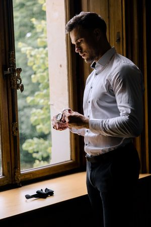 A person in a white dress shirt adjusts their cuff links while standing by a sunlit window.
