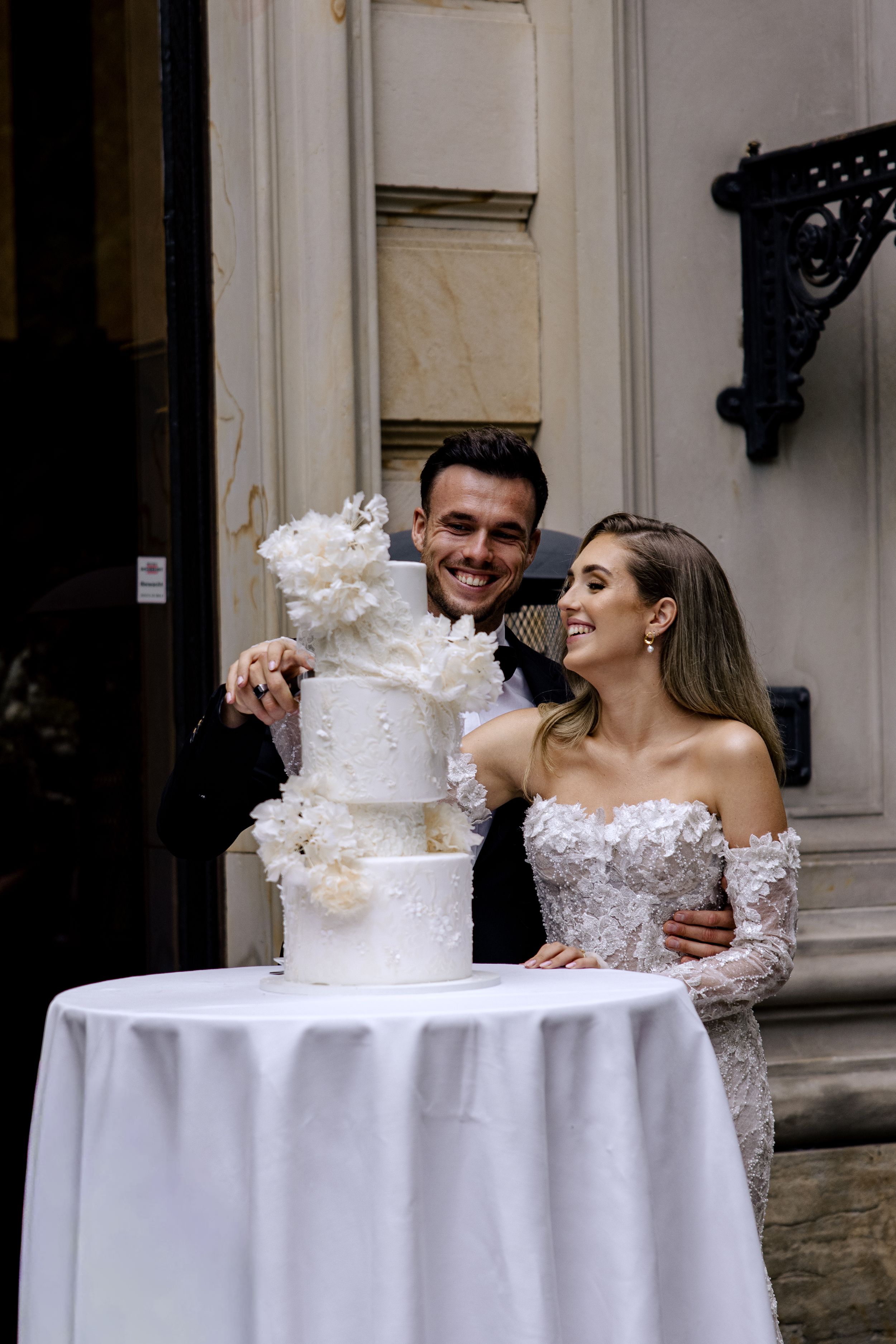 A couple celebrates at their wedding reception while cutting their elegant white tiered cake together.