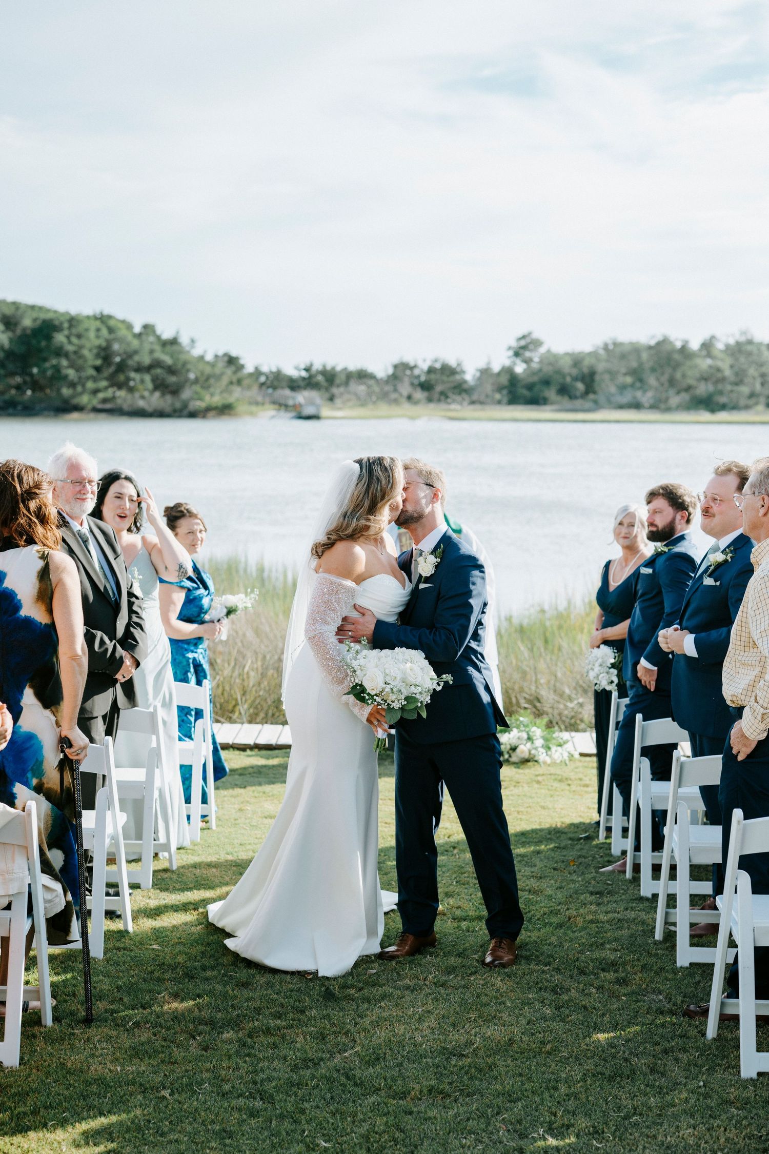 Outdoor waterfront wedding ceremony with white chairs and guests gathered on a grassy lawn overlooking a marina.