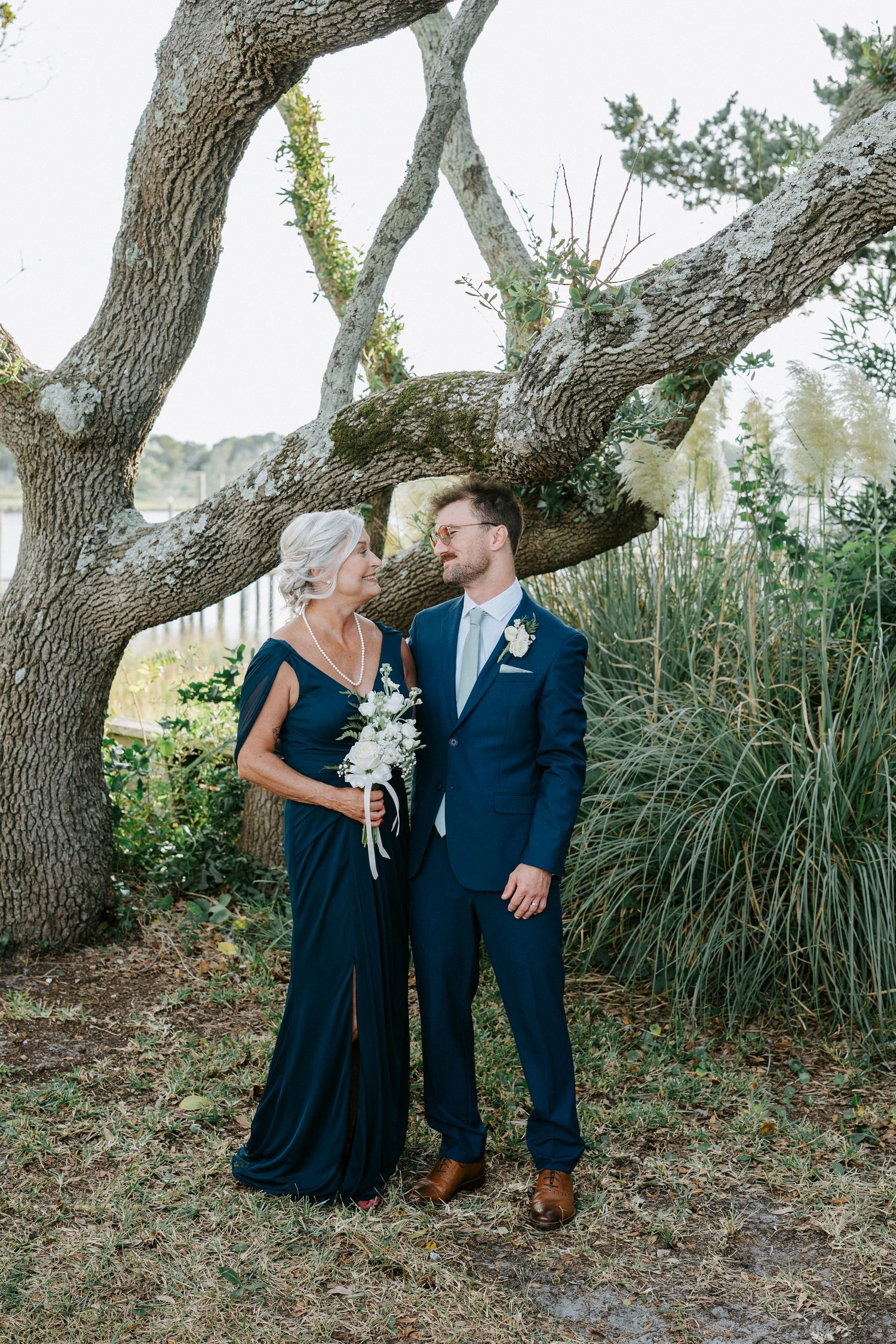 Couple in coordinating navy blue attire pose together under a gnarled tree branch in an outdoor setting.