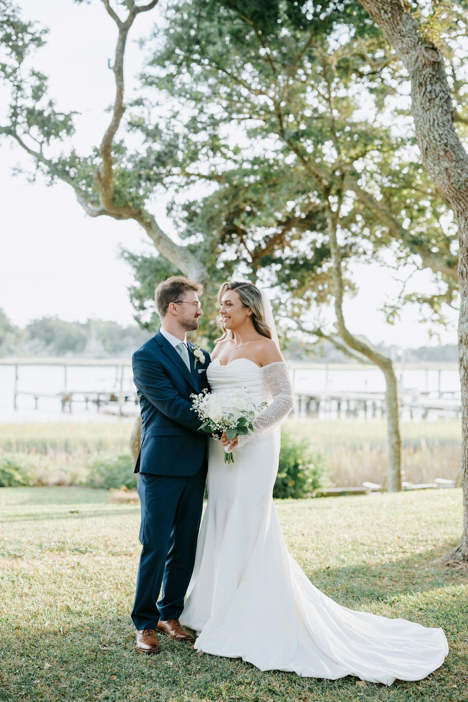 Elegant wedding couple shares intimate moment under oak trees with coastal marsh backdrop.