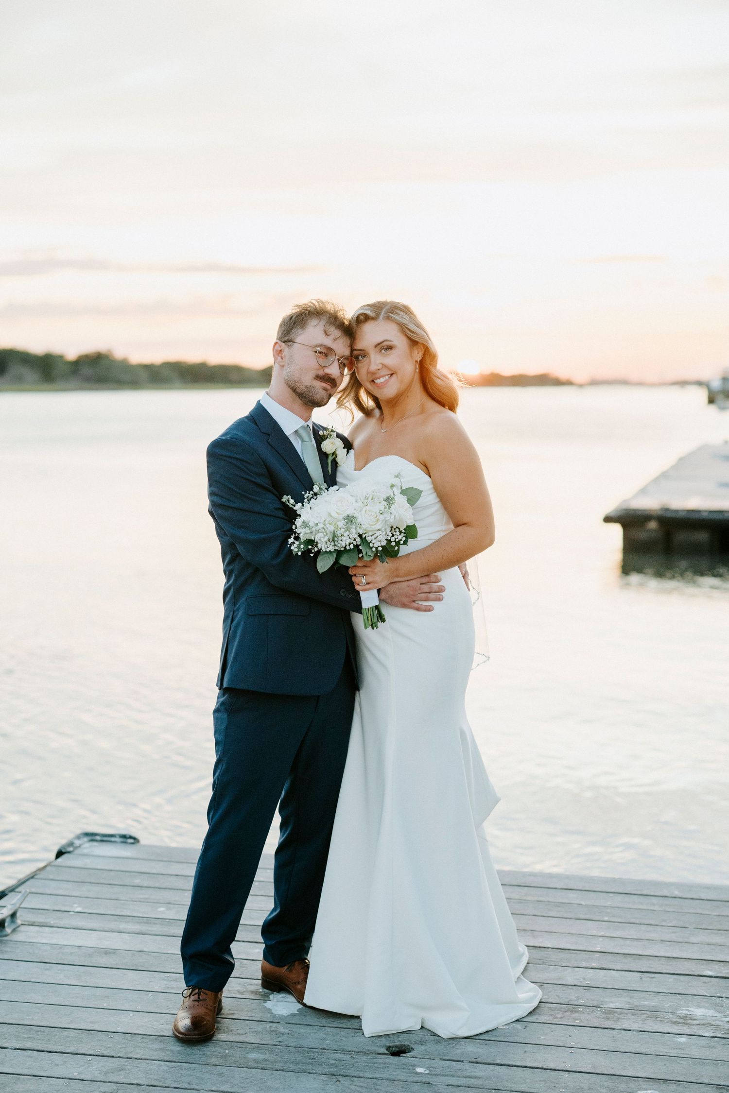 A couple in wedding attire shares an intimate moment at sunset on a wooden dock by the water.