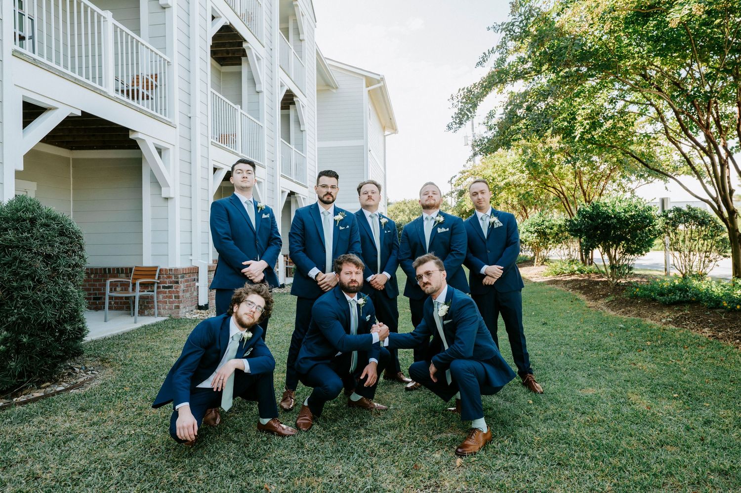 Groomsmen in navy suits pose casually on lawn in front of white building.