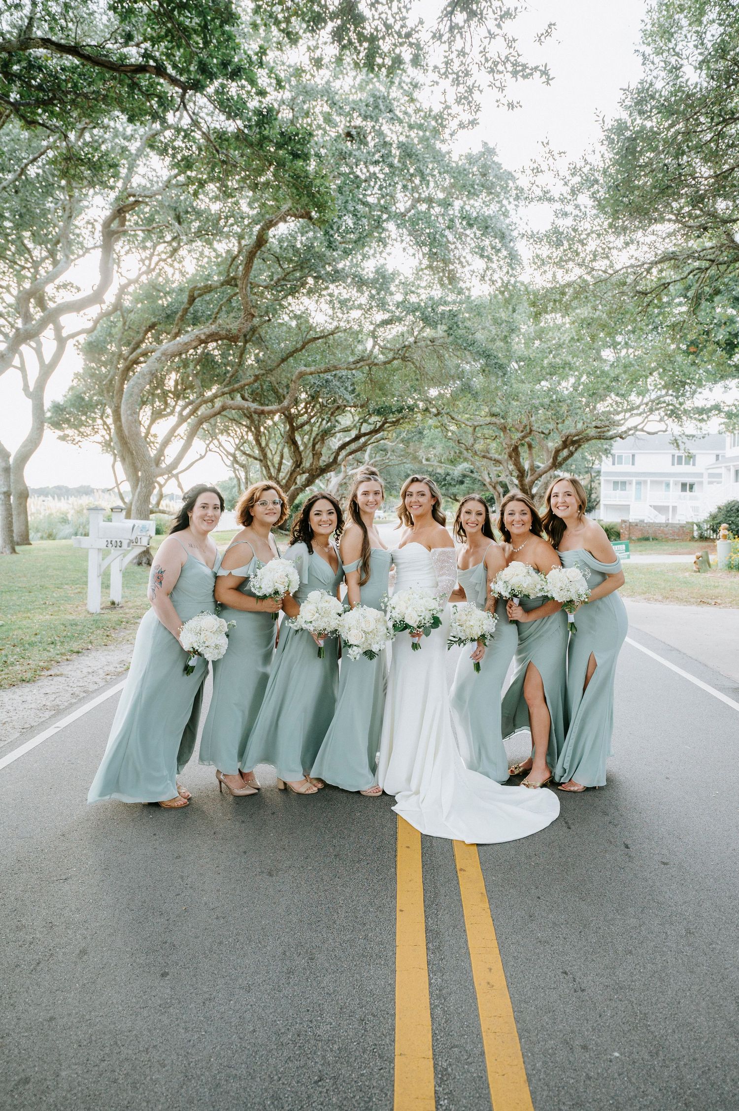 Bridesmaids in sage green dresses pose together casually on a tree-lined road with the bride in white.
