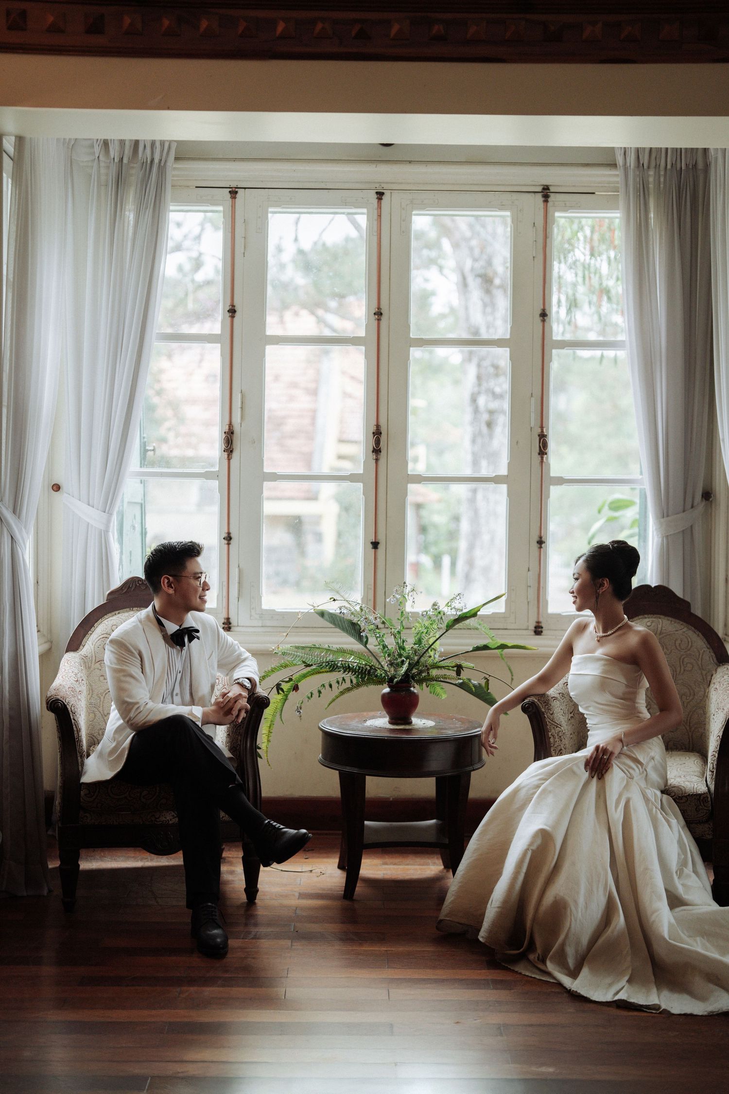 Bride and groom sit facing each other on antique chairs by large French windows in soft light.