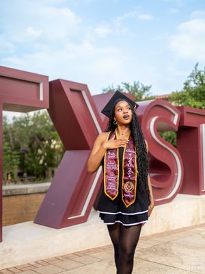 Graduate in cap and gown poses with decorative stoles against large outdoor university letters.