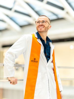 Graduate wearing orange Texas stole and white lab coat poses in bright indoor setting.