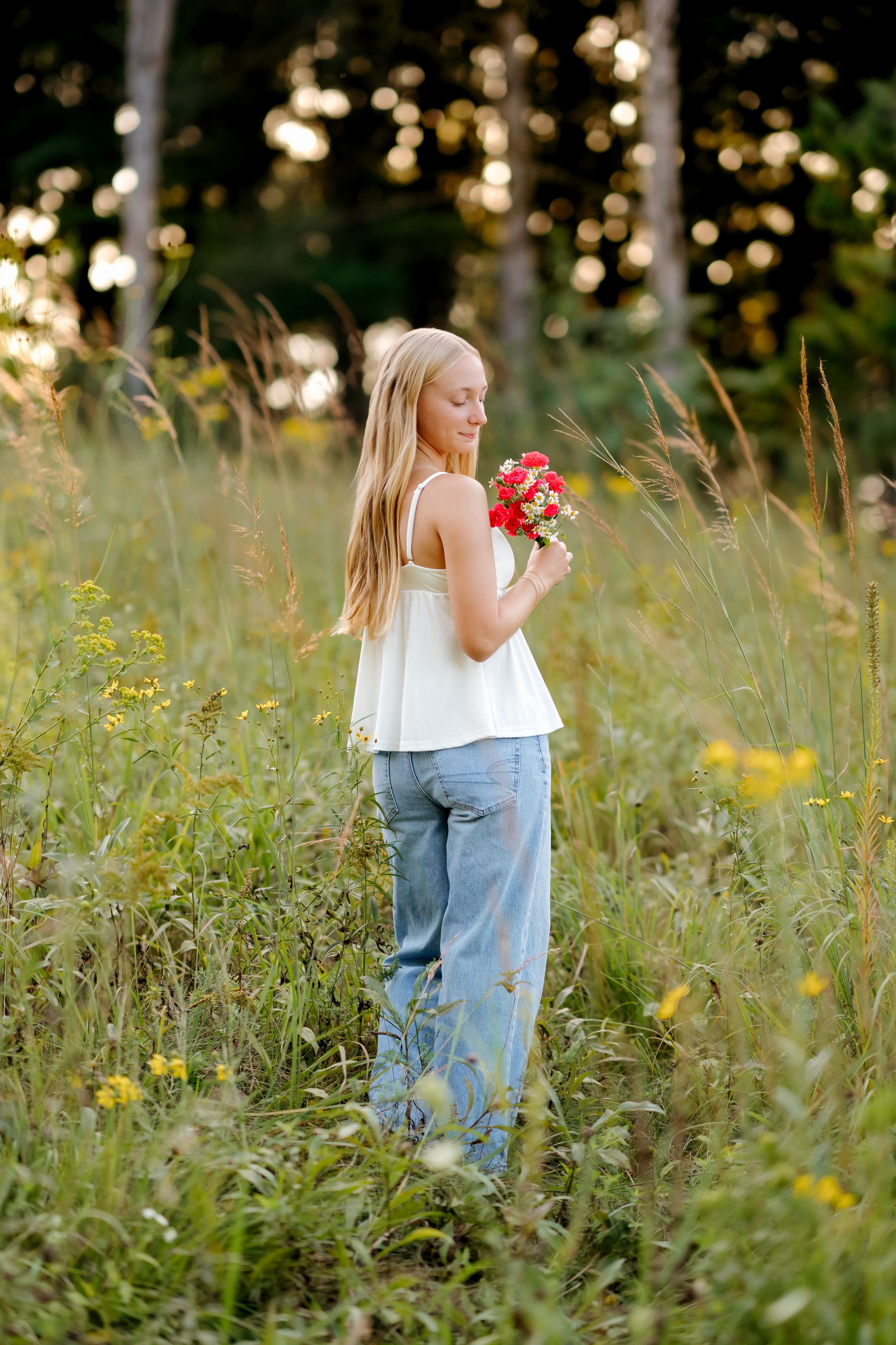 A young blonde girl in white top and jeans holds red flowers while standing in a lush green meadow at golden hour.