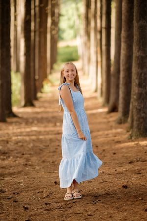 Smiling teenage girl in a light blue maxi dress walking through a pine tree forest path on a sunny day.