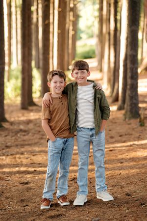 Two smiling boys hugging in a sunlit pine forest path, wearing casual jeans and jackets.