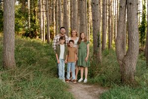 Family of five smiling together on a forest path surrounded by tall pine trees and lush green grass.