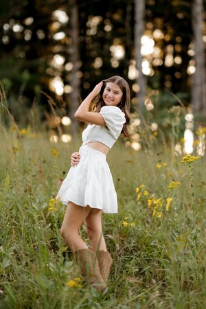 Young woman in white crop top and skirt standing in a wildflower meadow with forest bokeh background.