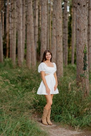 Young woman in white mini dress and cowboy boots posing among tall pine trees in a forest setting.