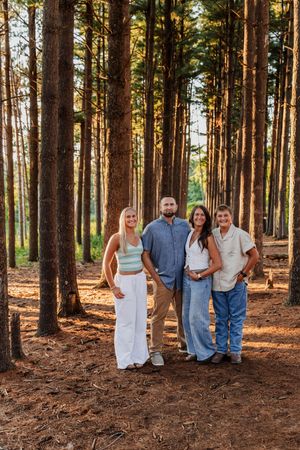 Four people smiling together in a sunlit pine forest, wearing casual neutral outfits for a family portrait.