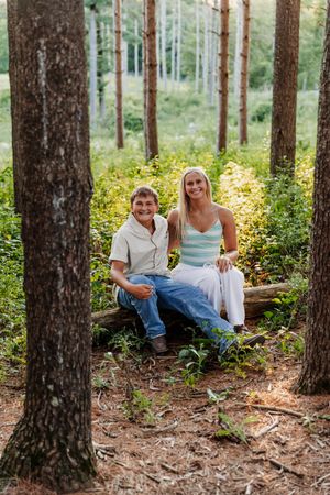 A couple smiles while sitting on a fallen log surrounded by tall pine trees and lush greenery in a forest.