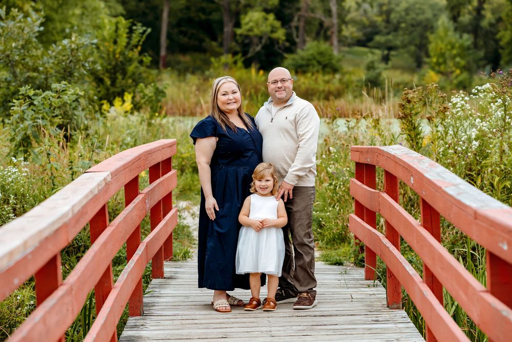 A couple shares a tender moment on a red wooden bridge surrounded by lush summer greenery and wildflowers.