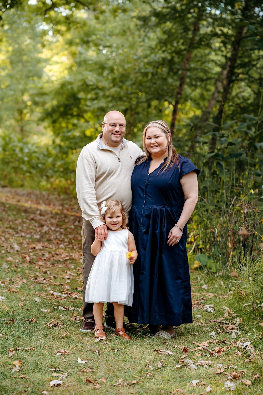 A family poses together outdoors in autumn, with fallen leaves on the ground and green trees in the background.
