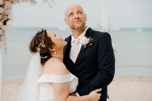 A couple in formal wedding attire shares a joyful laugh together on a beach.
