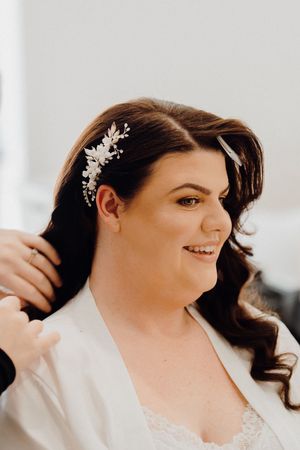 A bride getting ready on her wedding day wearing a white robe with curled hair adorned with delicate hair accessories.
