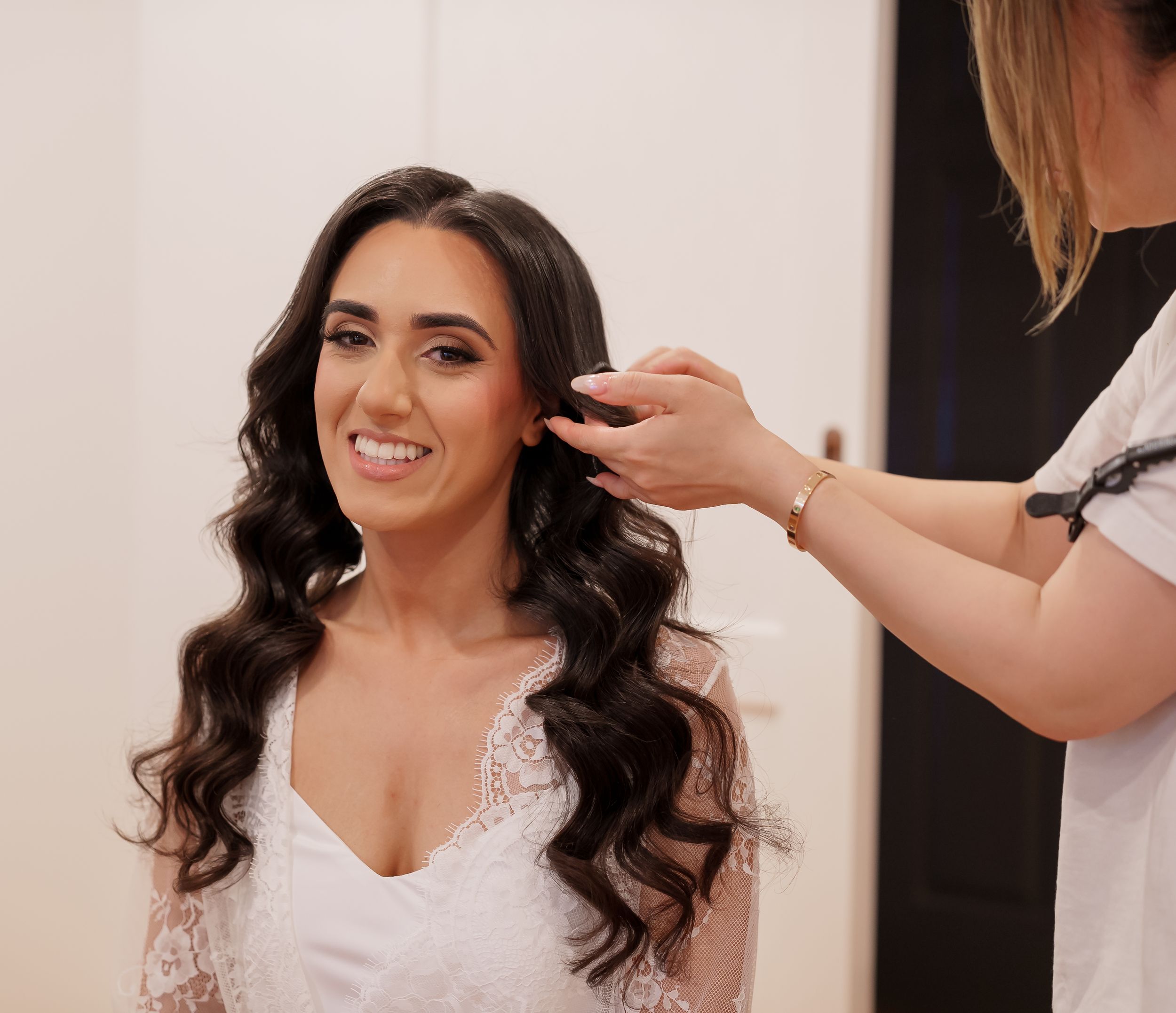 Wedding hairstylist arranging dark curled hair on bride wearing white dress during bridal preparation.