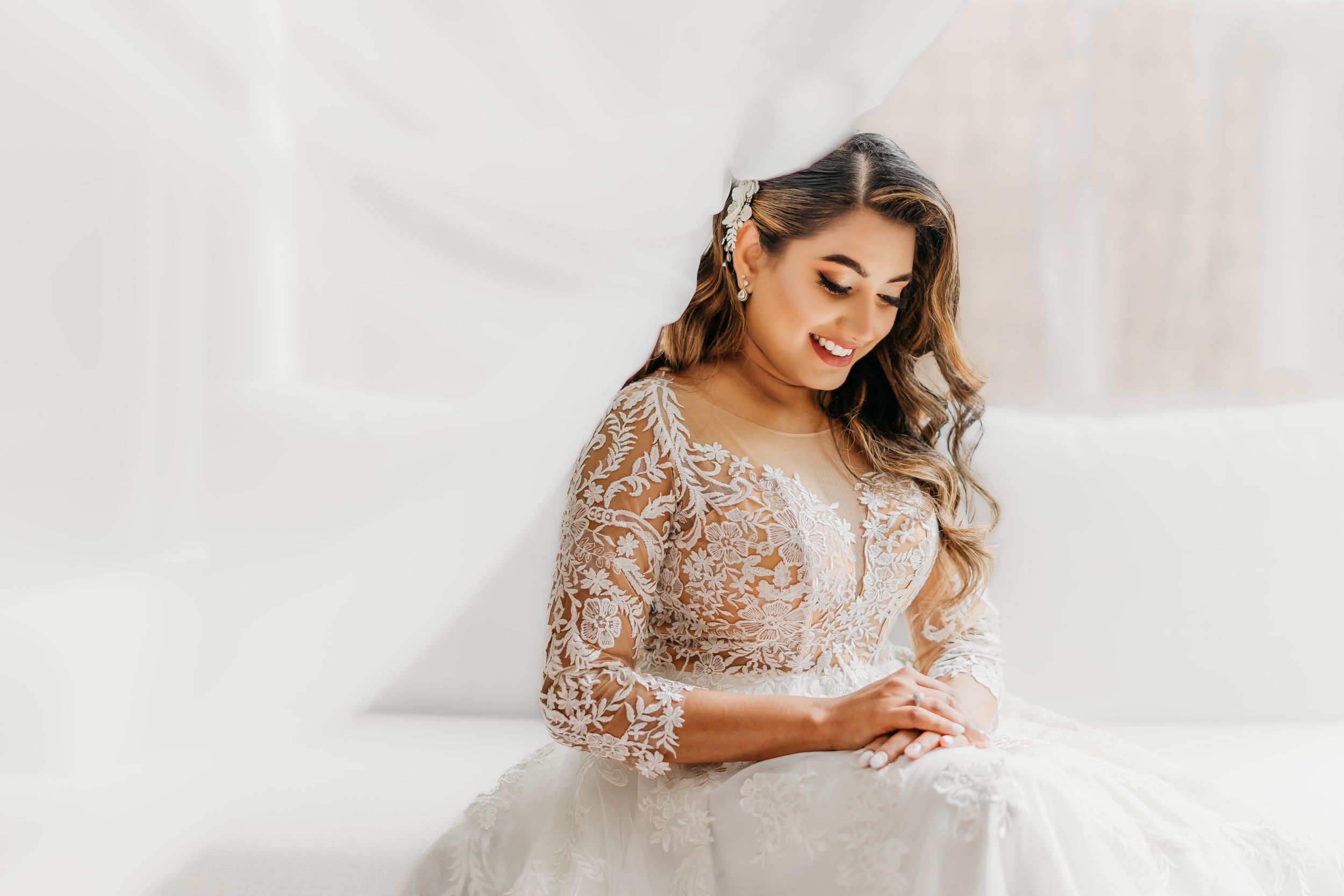 The bride sits elegantly in white lace bridal gown while looking down at her lap in ethereal wedding portrait.