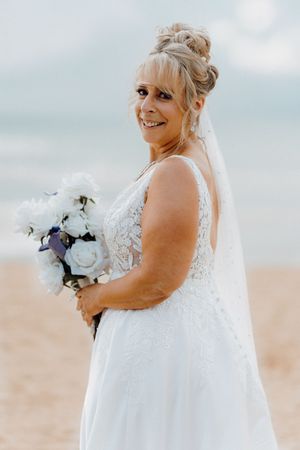 A bride in a flowing white lace wedding dress poses on a beach with a bouquet of white and blue flowers.