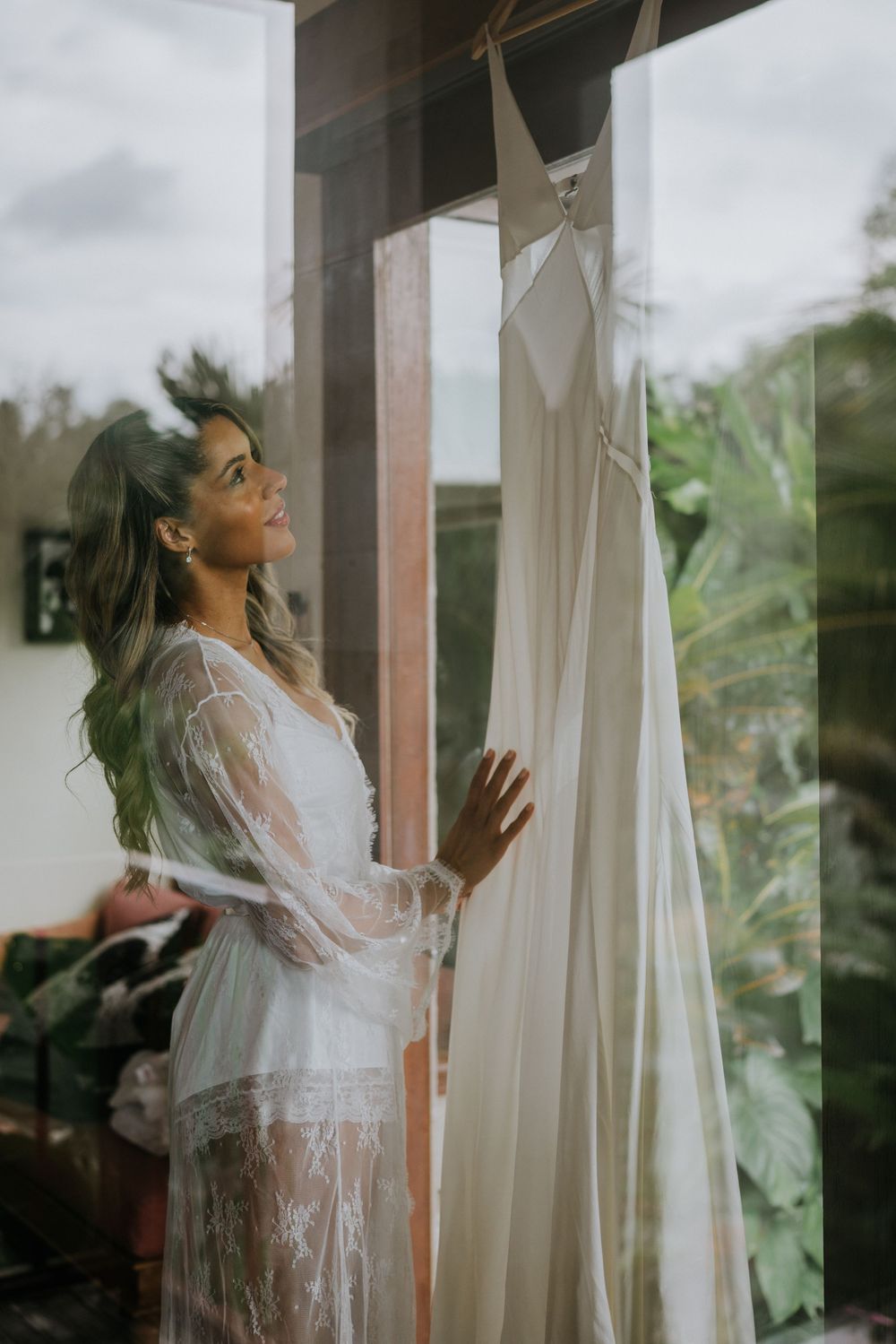 Bride in a white robe stands by sheer curtains in morning light during wedding preparations.