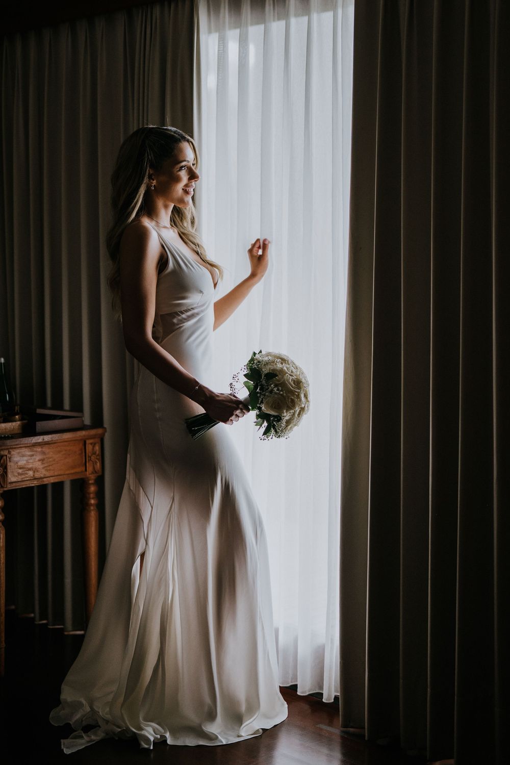 Silhouetted bride holding bouquet poses near window in dramatic lighting.
