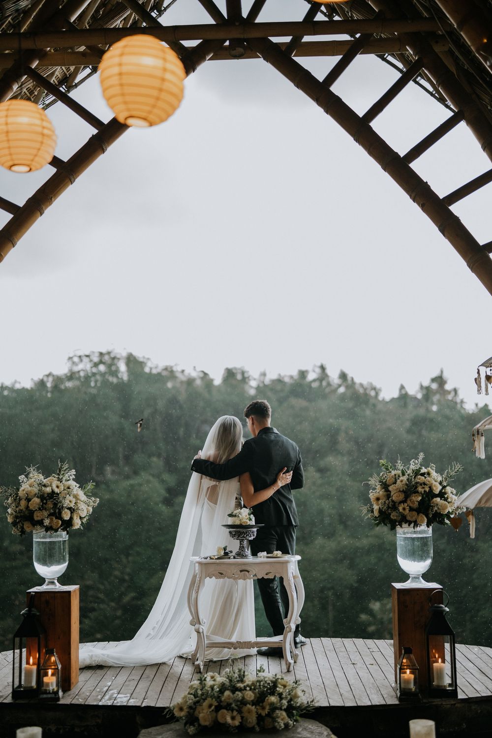 Wedding reception setup on wooden deck with orange lanterns and mountain view at dusk.