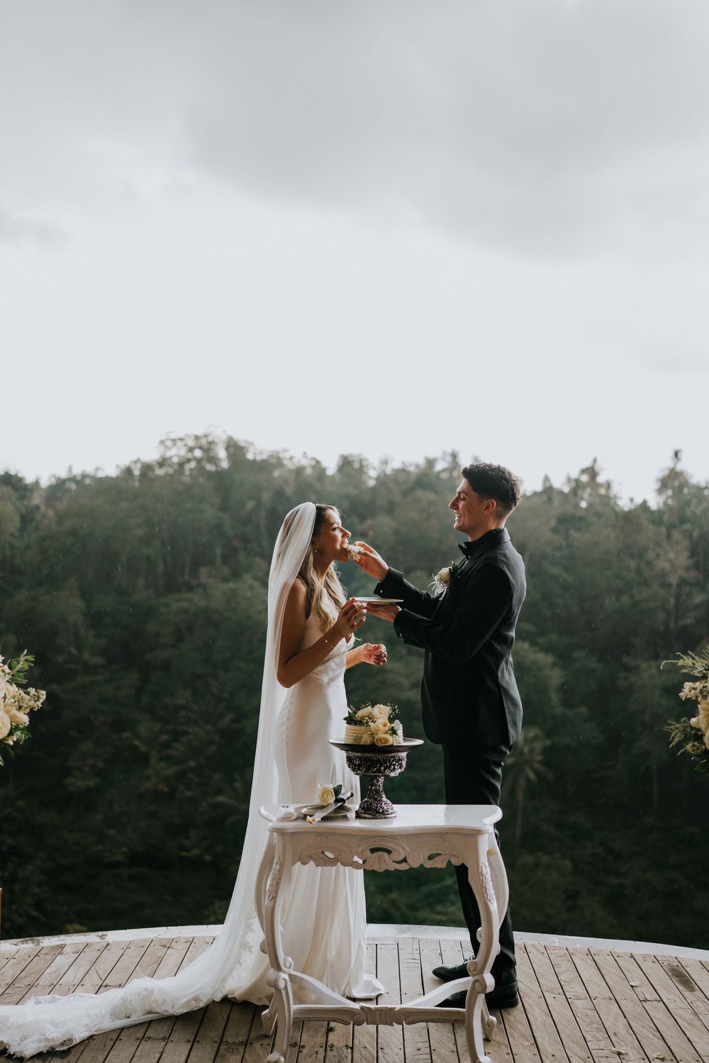 Newlyweds share cake cutting moment on outdoor deck with forest views.
