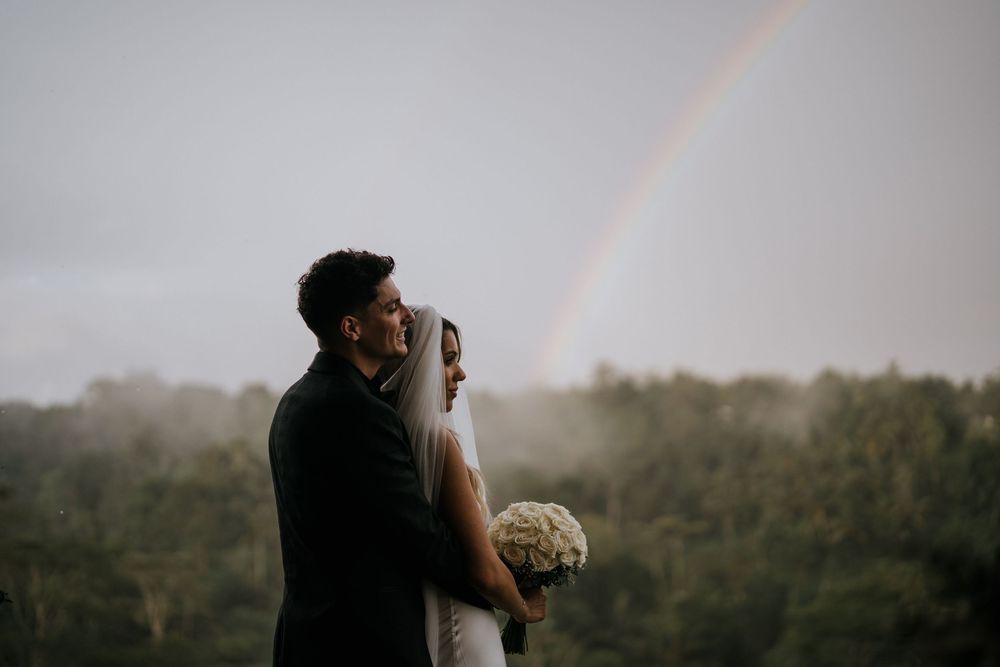 Silhouetted couple against dramatic sky with rainbow during sunset wedding photoshoot.