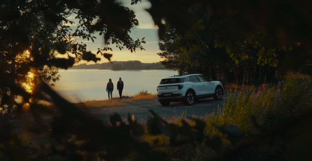 Silhouettes of people walking along a scenic mountain path at sunset with a parked car in the distance.