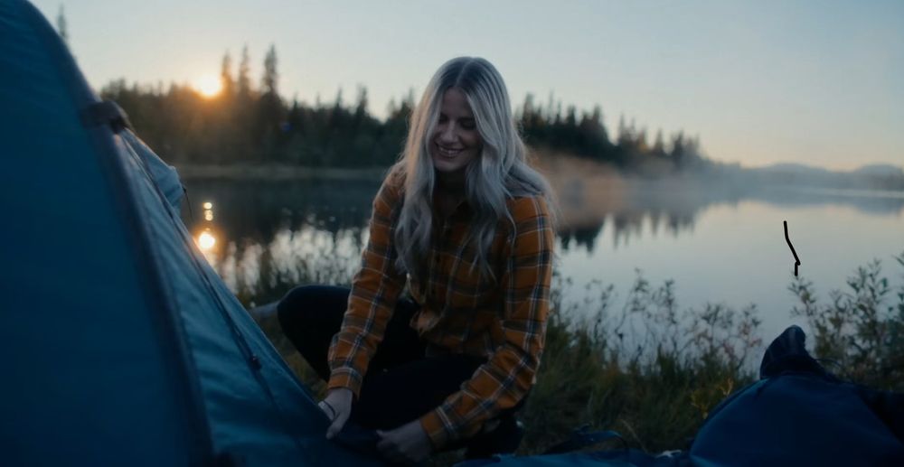 Campers gather by a lakeside at sunset with camping equipment and tents visible.