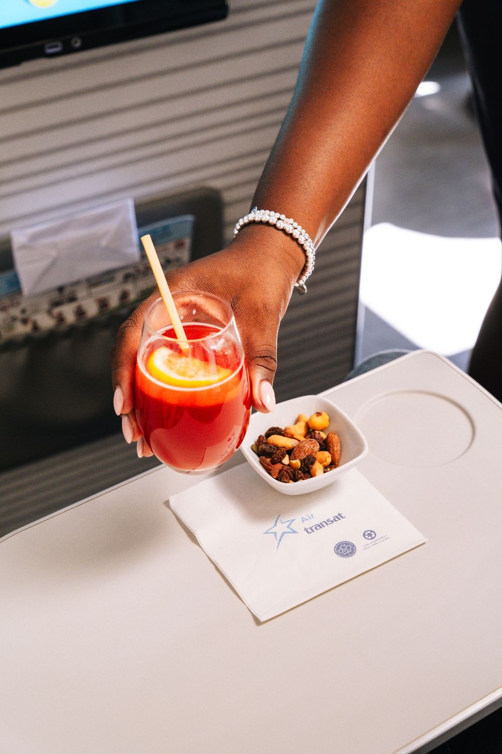 Red cocktail drink being served with a small bowl of mixed nuts and garnish on a white surface.