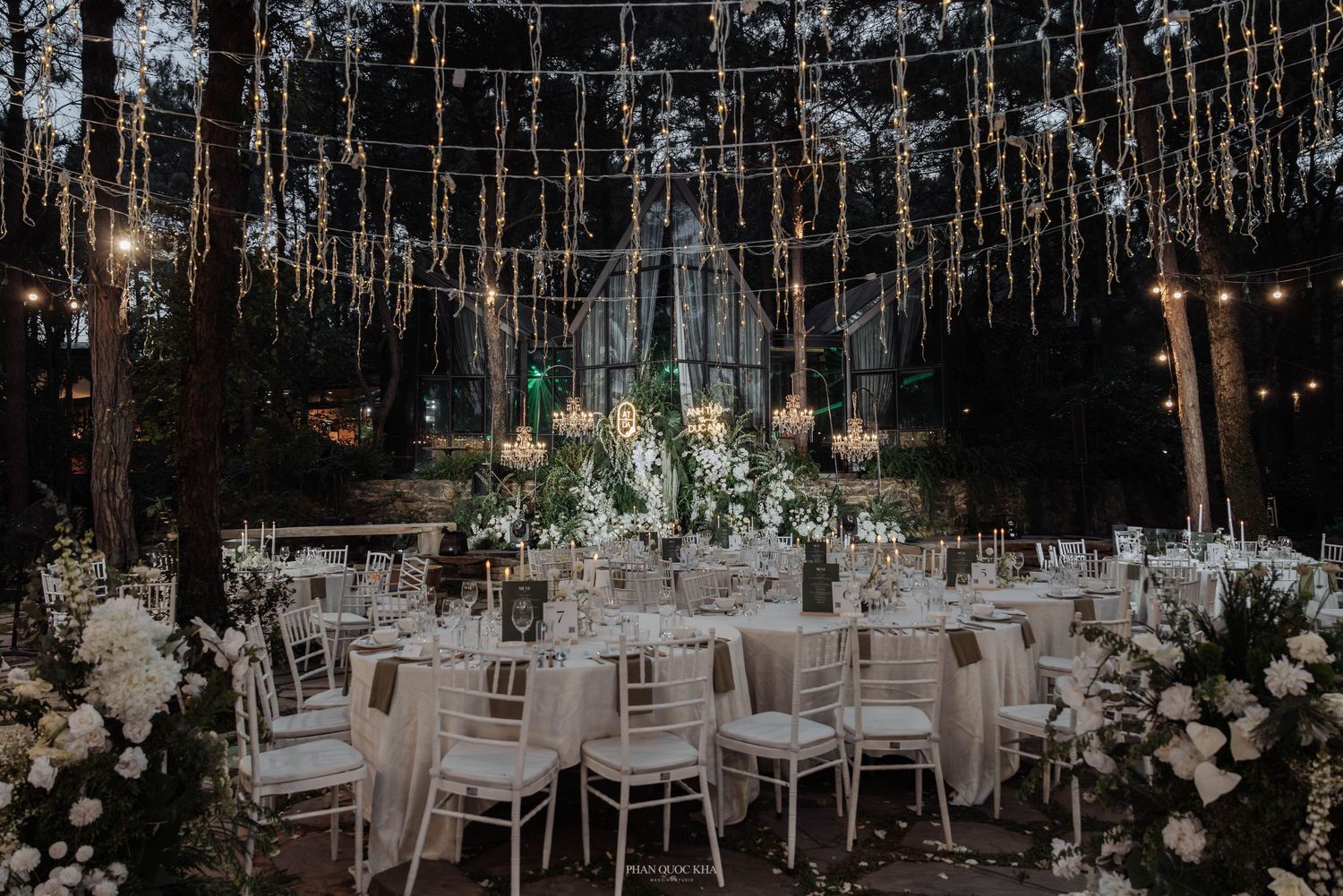 Enchanting forest wedding reception with fairy lights, chandeliers, white florals, and chiavari chairs.