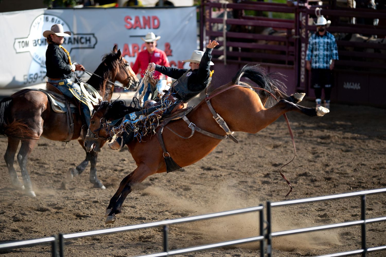 A cowboy rides a bucking bronco during a rodeo competition in a dirt arena with spectators watching from behind fencing.