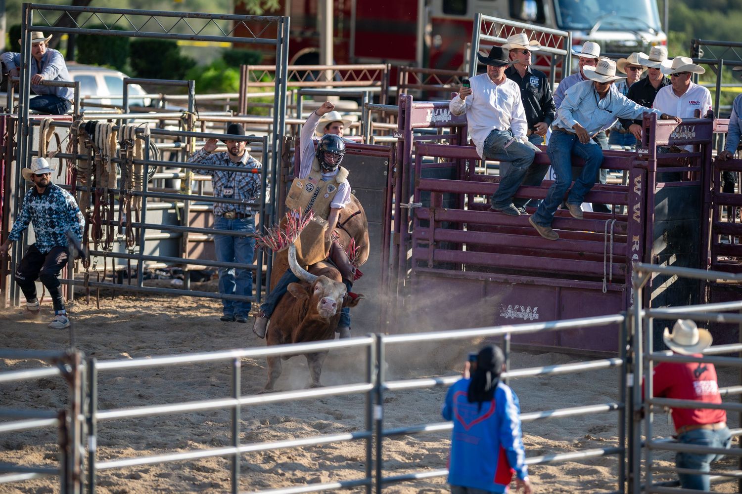 Competitive rodeo action at a western arena shows cowboys and spectators gathered around metal gates and fencing.