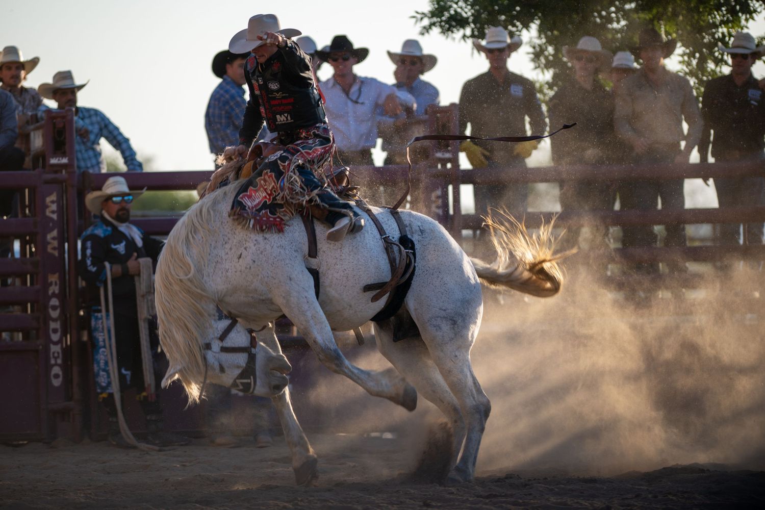A cowboy rides a bucking horse in a rodeo arena while spectators watch from the fence in dusty conditions.
