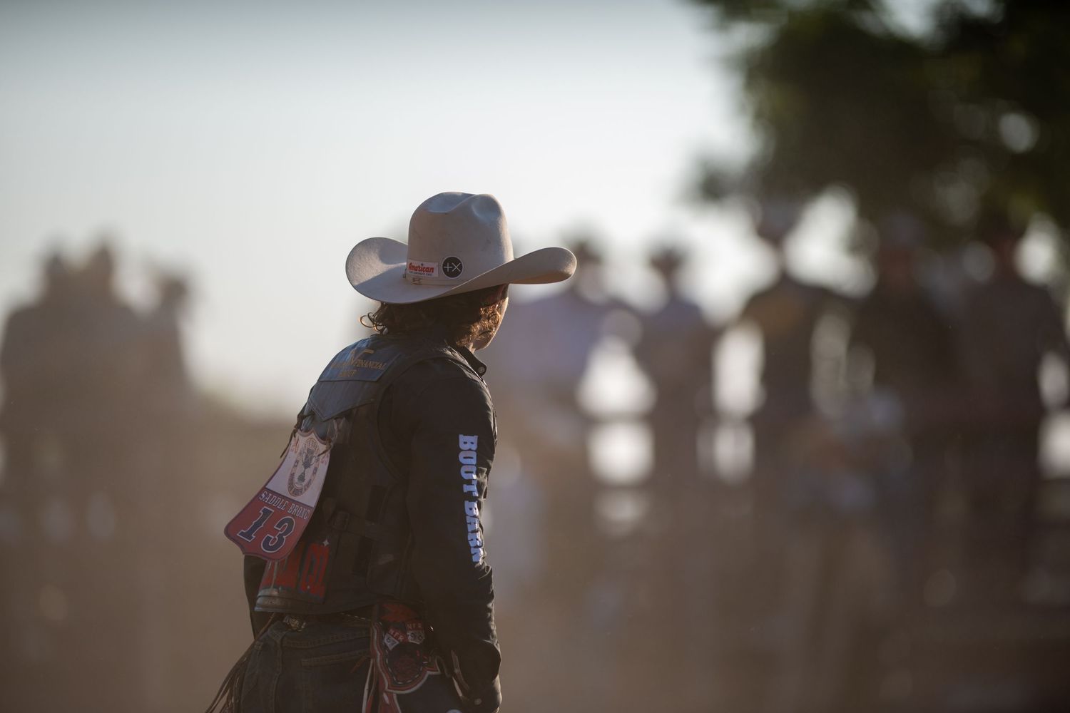 A cowboy rides a bucking bronco in silhouette against a dusty sunset at a rodeo competition.