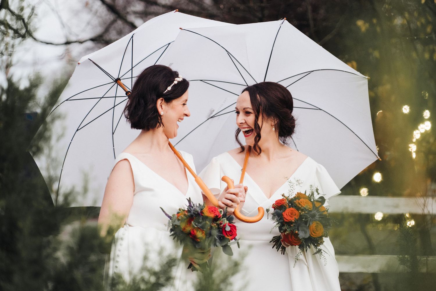 Two brides share a romantic moment under a white umbrella during an outdoor wedding photoshoot.