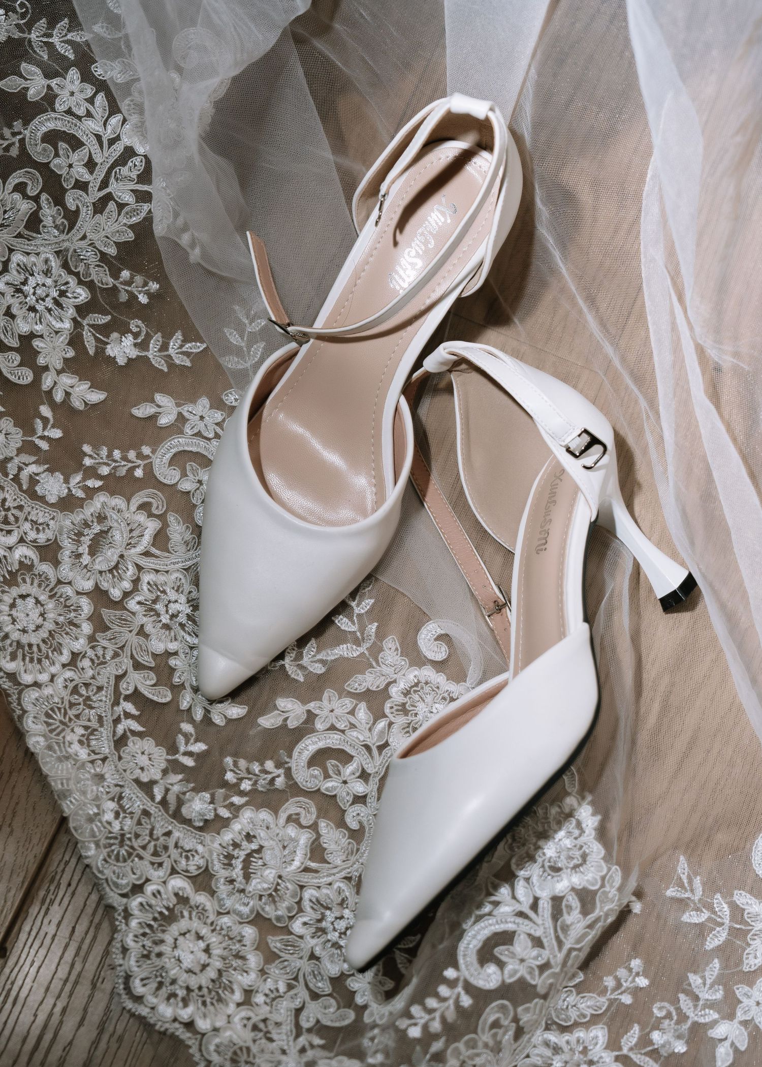 White pointed-toe bridal heels with ankle strap resting on lace wedding veil on wooden floor.