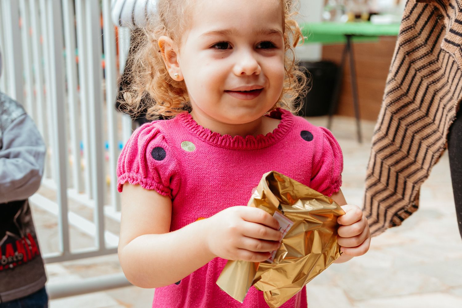 A child in a bright pink dress smiles while holding a crumpled brown paper bag.