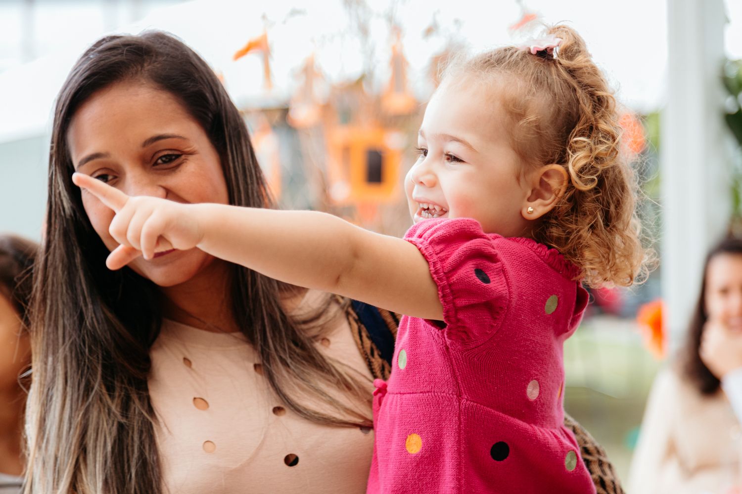 A child in a pink polka dot dress points excitedly while being held during an outdoor gathering.