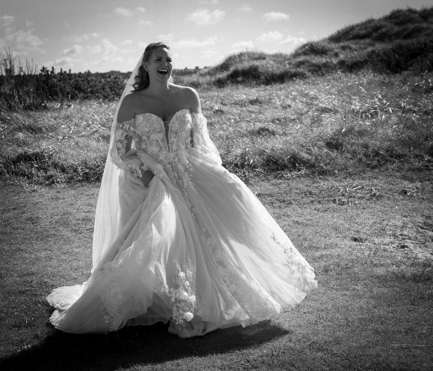 Black and white photo of radiant bride in strapless lace wedding gown walking outdoors on grassy hill.