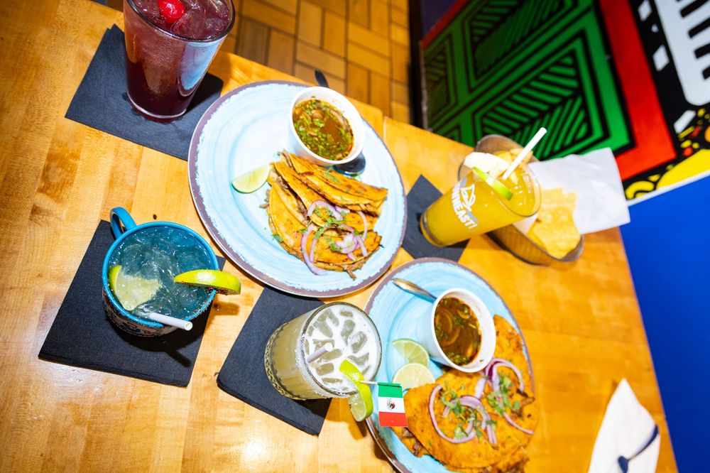 Overhead view of Mexican restaurant table with tacos, guacamole, chips, and colorful drinks on a wooden surface.
