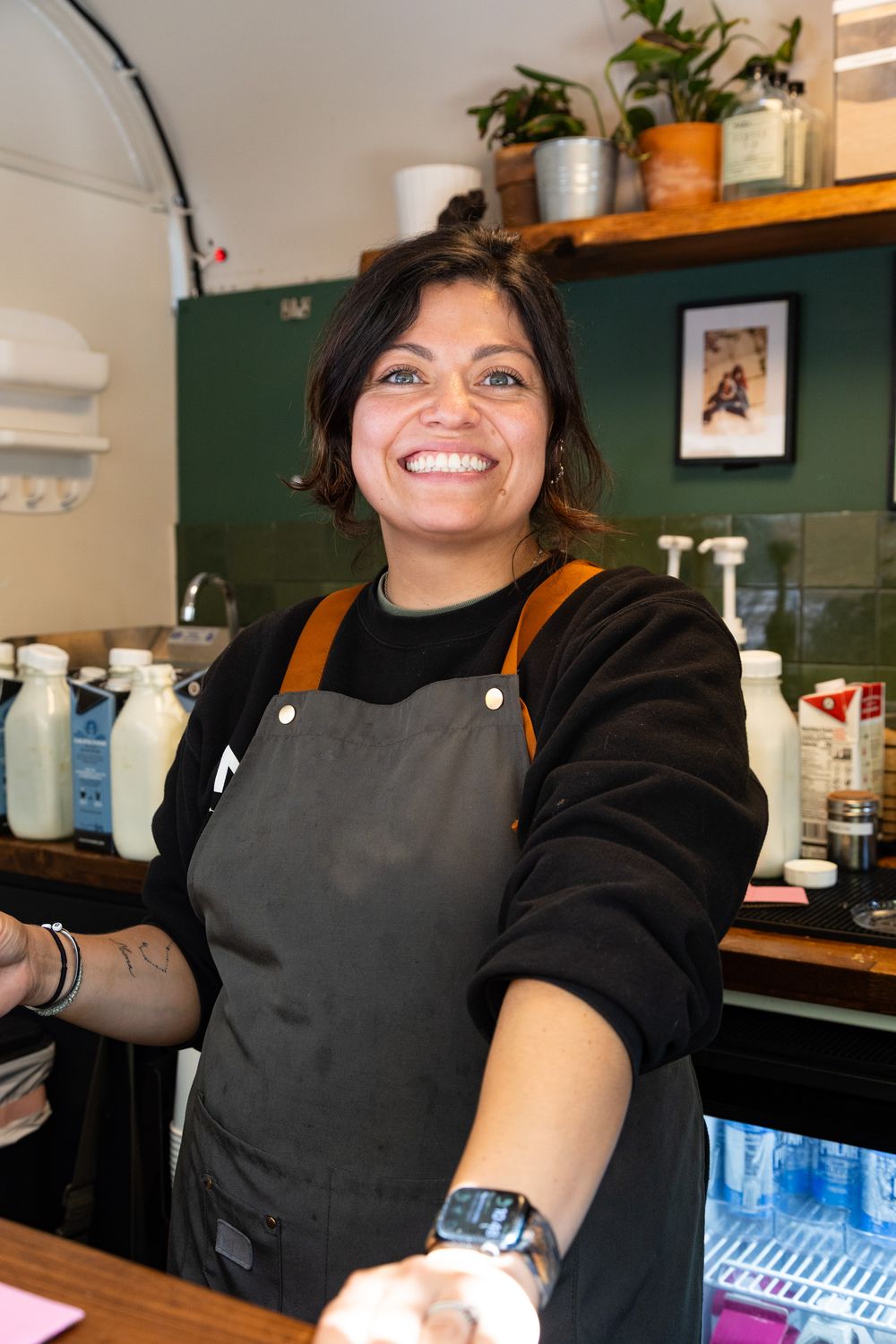 Barista wearing black shirt and gray apron smiles while working behind counter in coffee shop.