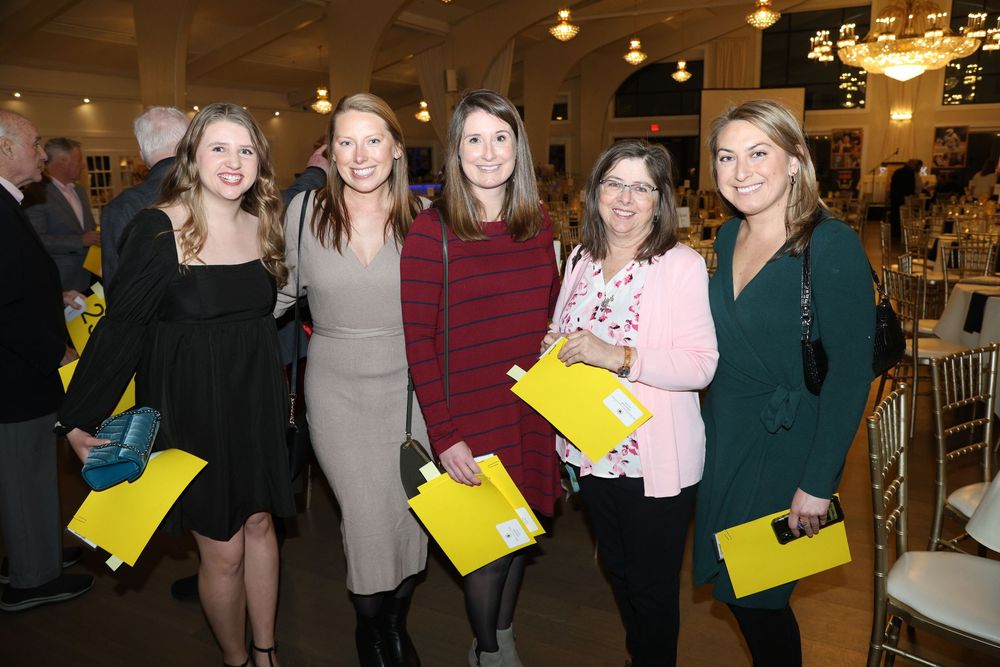 A group of friends holding yellow folders gather at an elegant indoor event venue with chandelier lighting.